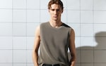 Placeholder for: Man in sleeveless shirt and dark pants stands against a tiled wall, hands in pockets, with soft lighting casting shadows.