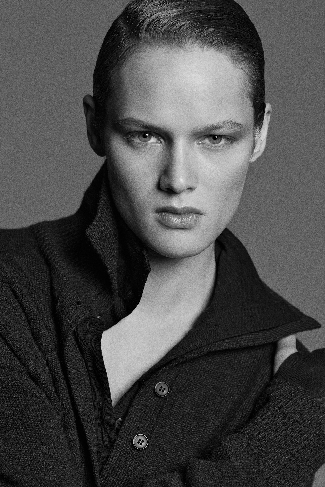 A black and white portrait of a person with slicked-back hair, wearing a dark buttoned cardigan with a collared shirt underneath, exuding a serious expression.