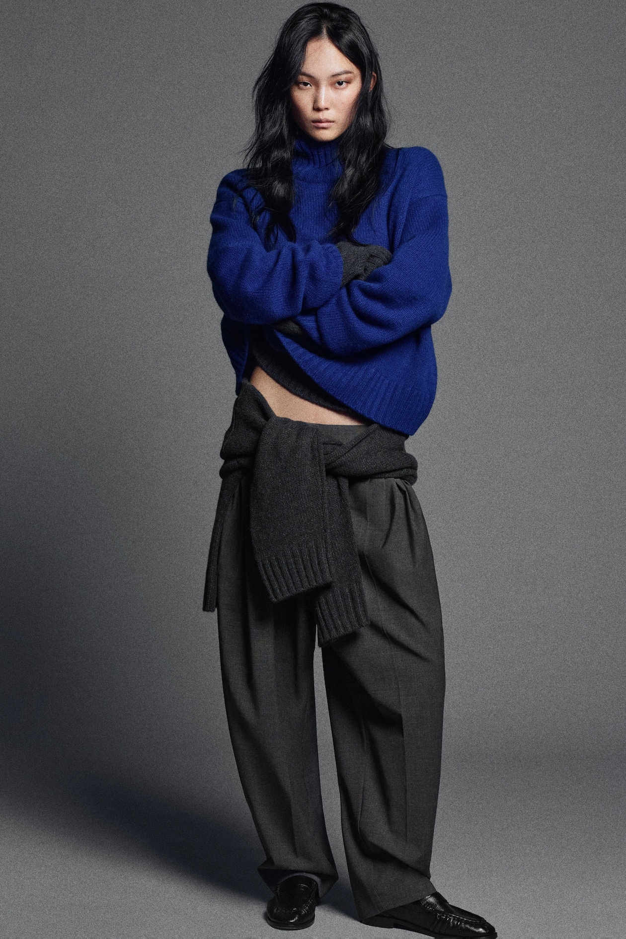 Person in a blue sweater with dark pants and a sweater tied at the waist, standing against a gray background.