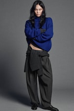 Placeholder for: Person in a blue sweater with dark pants and a sweater tied at the waist, standing against a gray background.