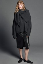 Placeholder for: A person wearing a dark gray oversized sweater, layered with a draped scarf, a black leather pencil skirt, and black slip-on shoes on a gray background.
