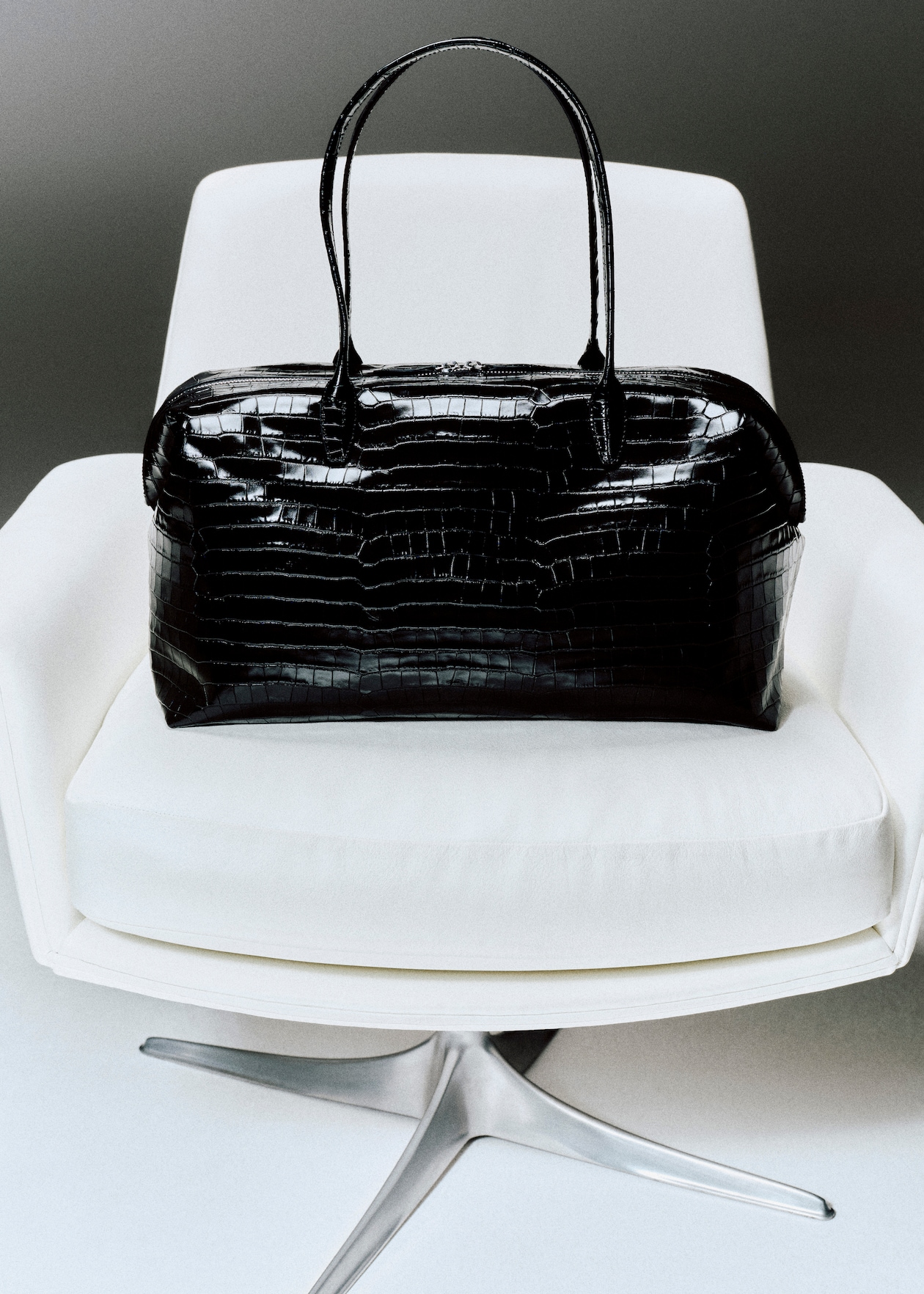 A black leather handbag with a glossy crocodile pattern rests on a modern white swivel chair.