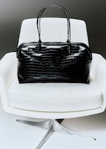 Placeholder for: A black leather handbag with a glossy crocodile pattern rests on a modern white swivel chair.