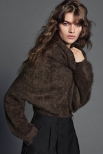 Placeholder for: A person with long wavy hair wearing a thick brown sweater and black pants, standing in front of a gray background, looking serious.