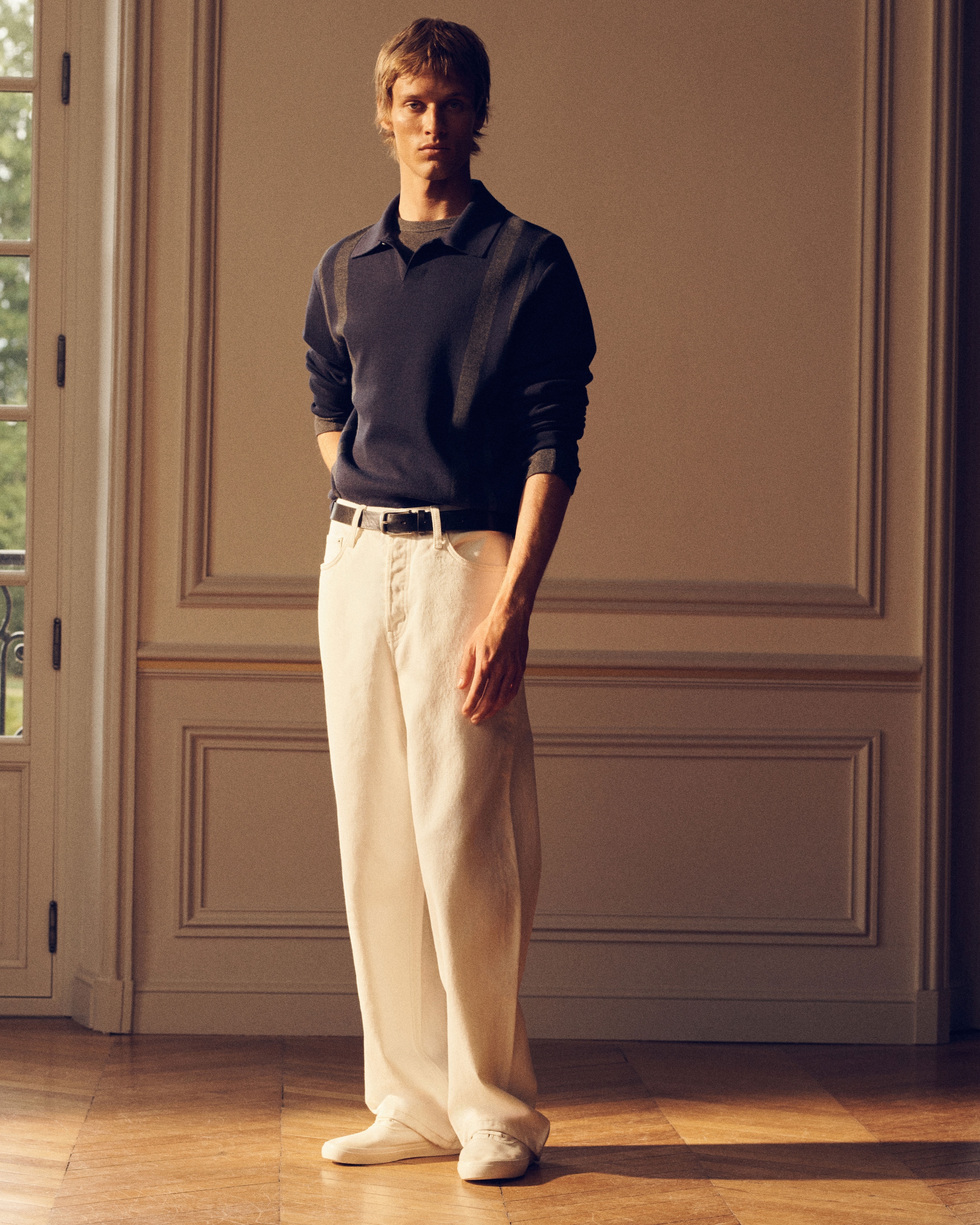 A person stands in a room with elegant paneling, wearing a dark long-sleeve shirt and light trousers, with hands in pockets, looking forward.