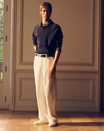 Placeholder for: A person stands in a room with elegant paneling, wearing a dark long-sleeve shirt and light trousers, with hands in pockets, looking forward.