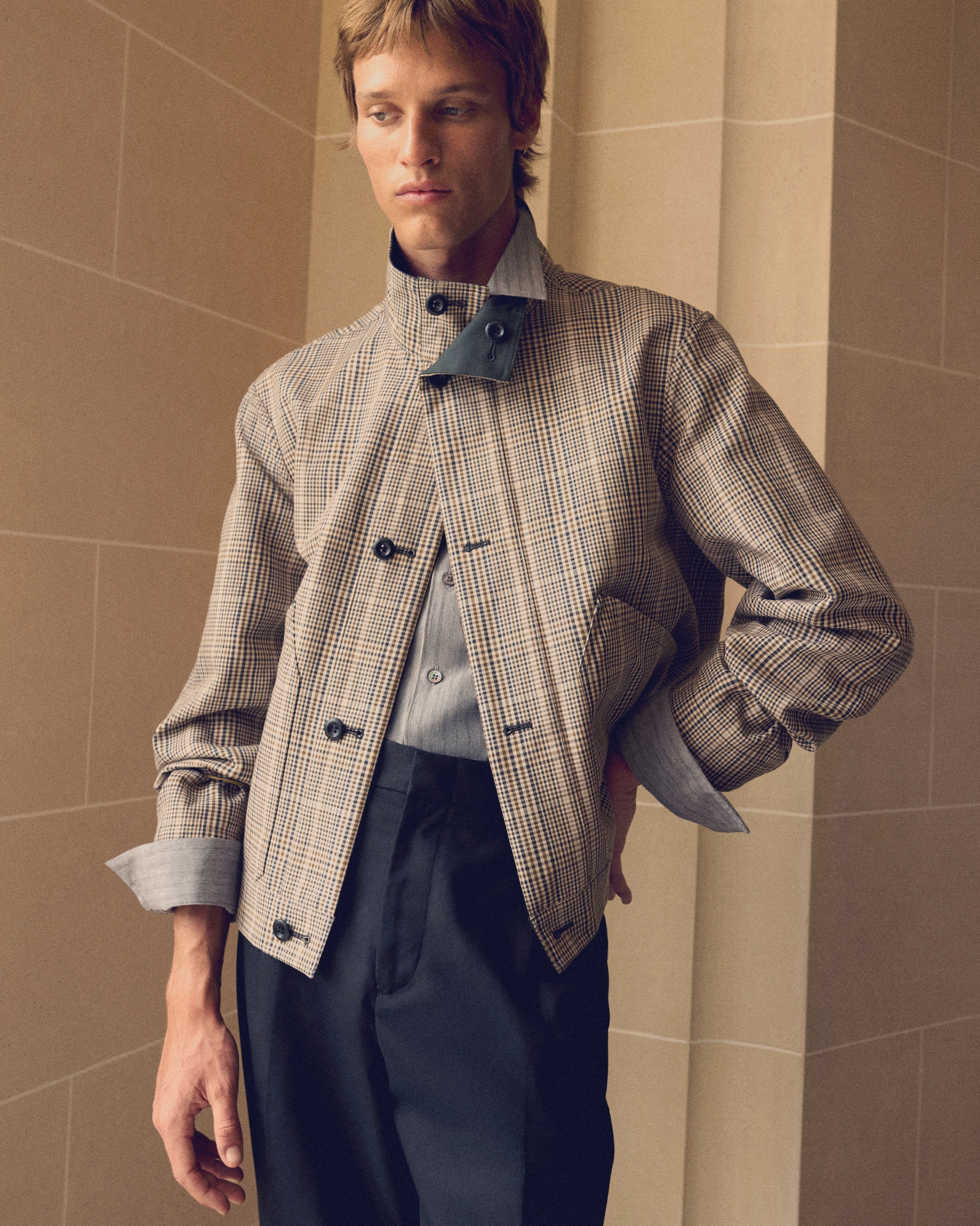 Person wearing a checkered jacket with a high collar, blue shirt, and dark pants, posing in front of a beige, tiled background.