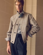Placeholder for: Person wearing a checkered jacket with a high collar, blue shirt, and dark pants, posing in front of a beige, tiled background.