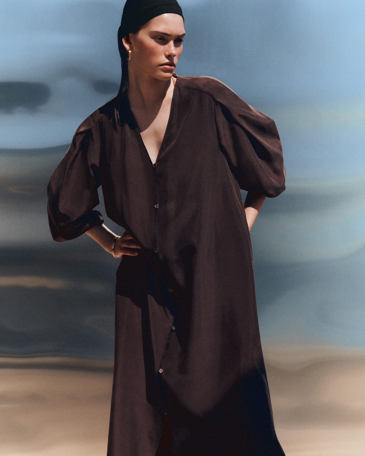 Person in a loose, dark brown dress with voluminous sleeves and a black headscarf, standing against a blurred outdoor background.