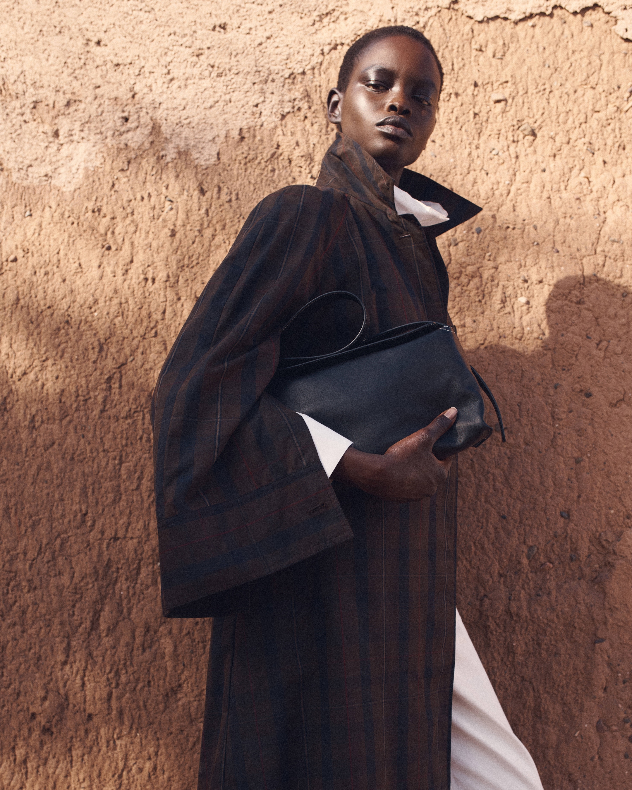 Fashionable person in a brown plaid coat, holding a black clutch, poses against a textured, earthy wall.