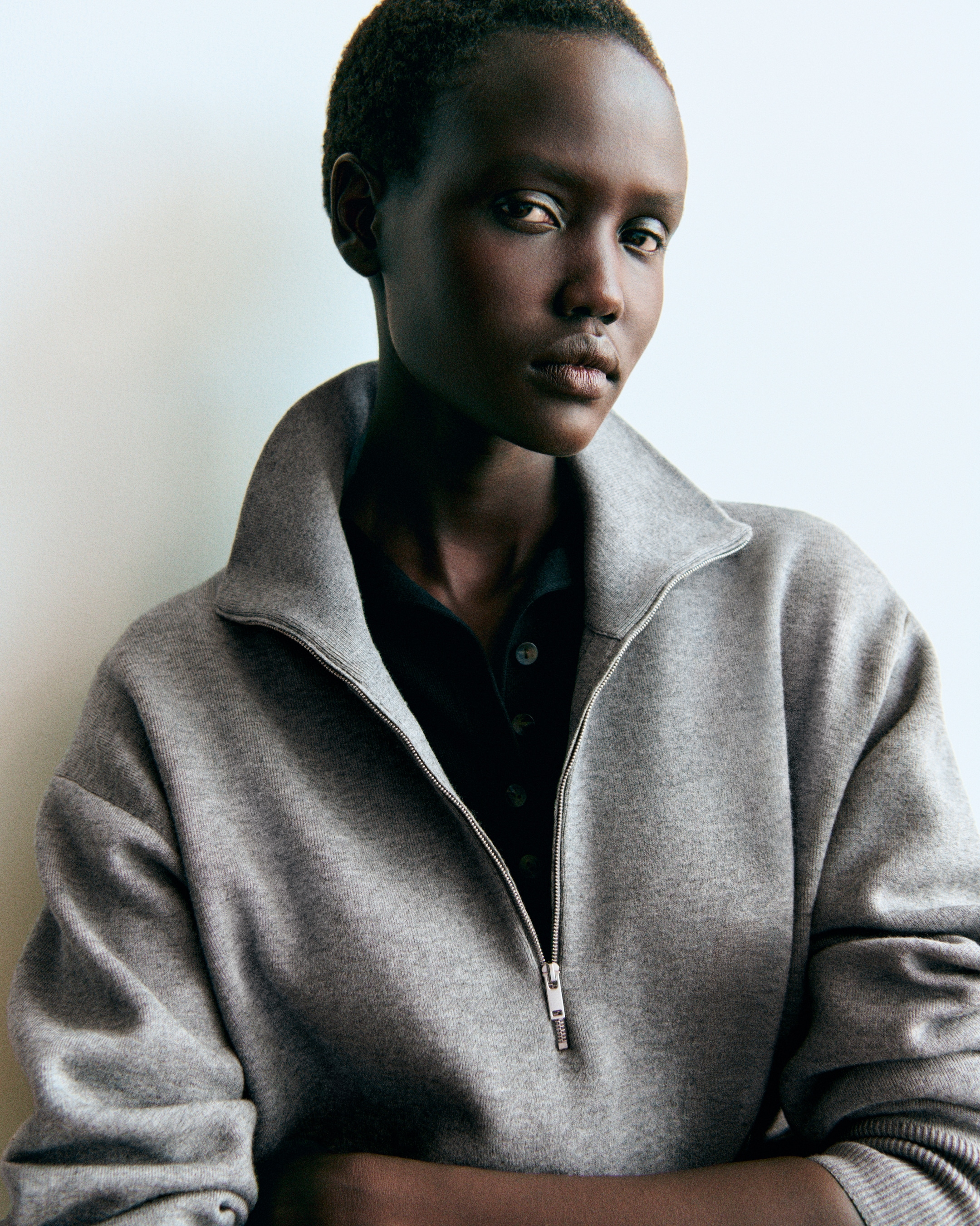 Person wearing a gray zip-up sweater over a dark shirt, standing against a light background.