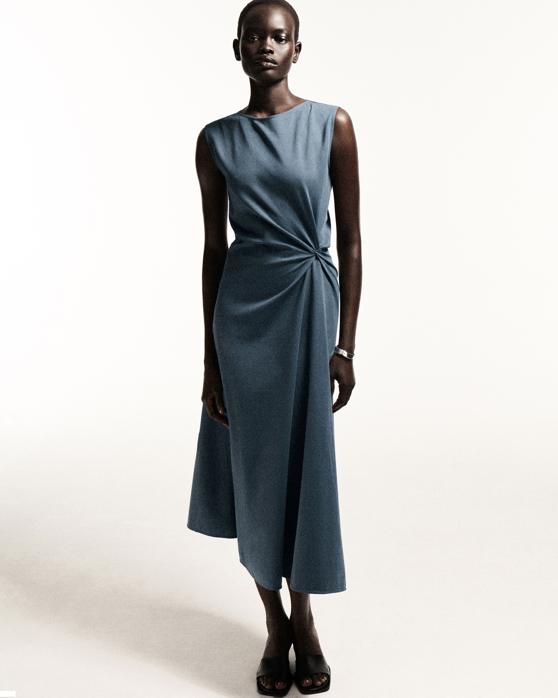 A person models a sleeveless, draped, blue dress with a twisted detail at the waist, standing against a plain white background.