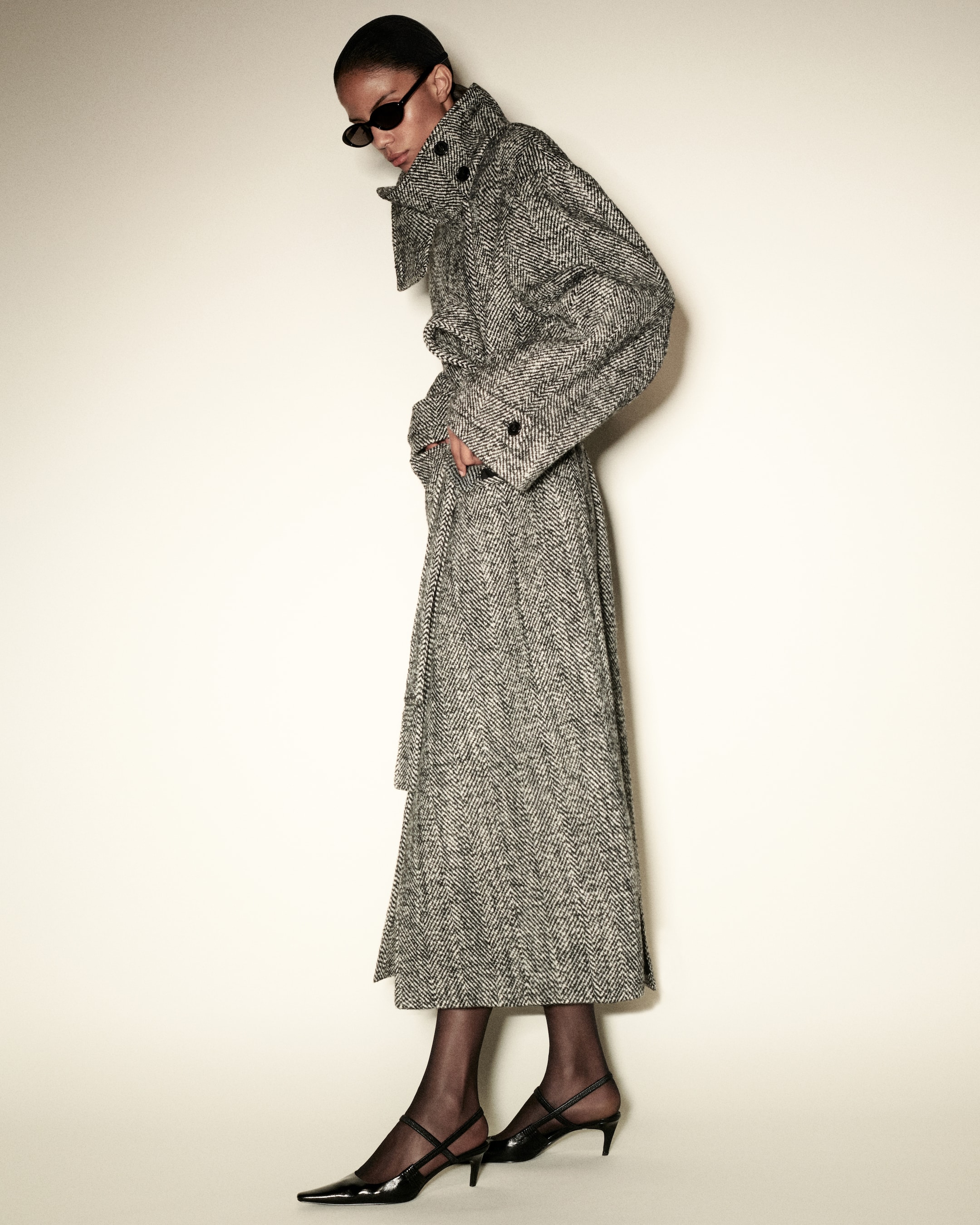 Person wearing a long herringbone coat, dark sunglasses, and black heels, standing against a plain background with a side profile view.