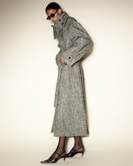 Placeholder for: Person wearing a long herringbone coat, dark sunglasses, and black heels, standing against a plain background with a side profile view.