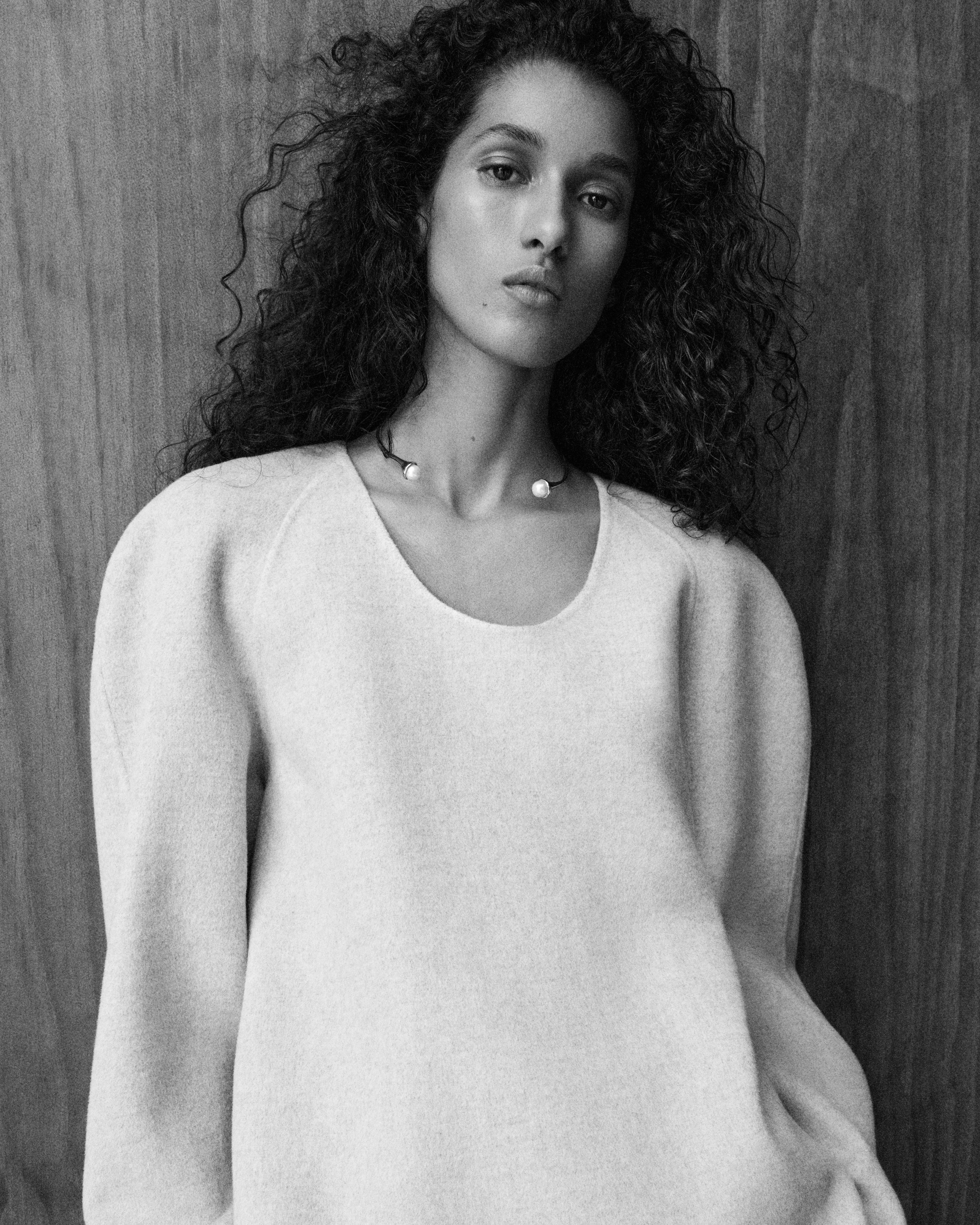 Woman with curly hair in a textured sweater poses against a wooden backdrop, exuding a calm and confident expression. Black and white.