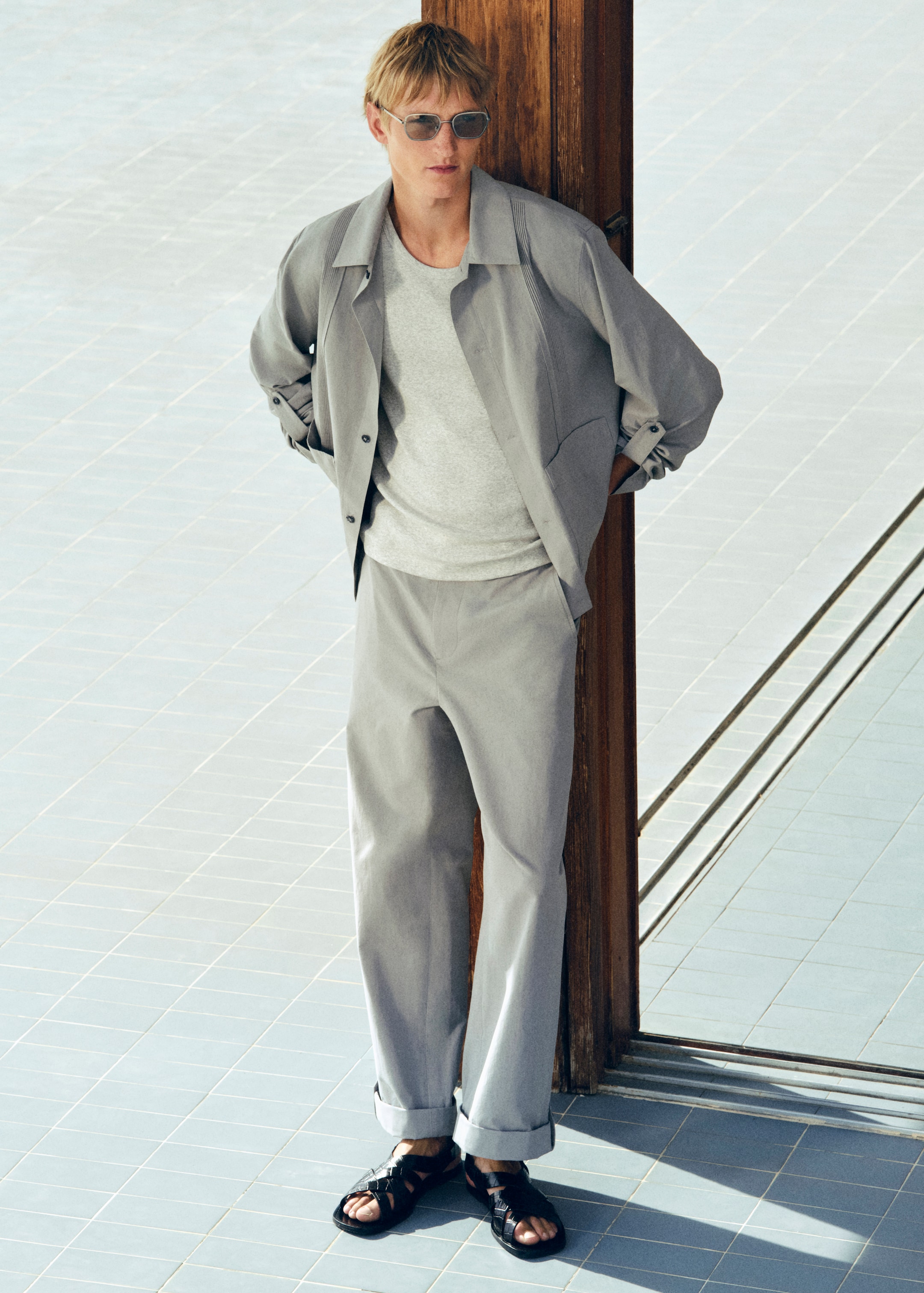 A person wearing a light gray outfit, sunglasses, and black sandals leans casually against a wooden pillar on a tiled floor.