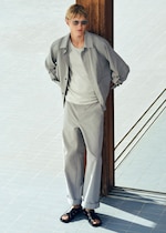Placeholder for: A person wearing a light gray outfit, sunglasses, and black sandals leans casually against a wooden pillar on a tiled floor.