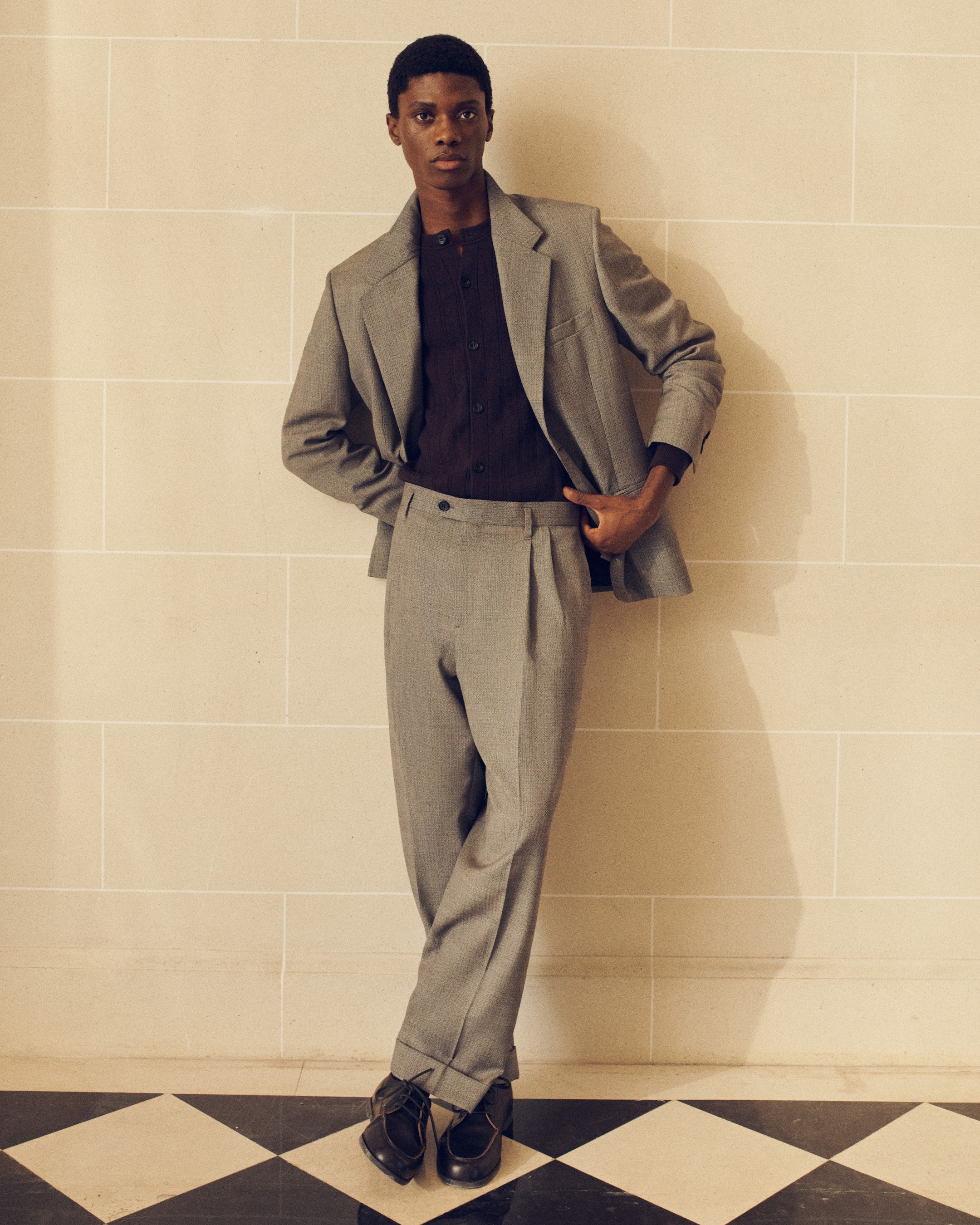Person in a gray suit and dark shirt poses against a tiled wall with one hand in pocket, standing on a black and white checkered floor.