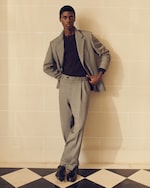Placeholder for: Person in a gray suit and dark shirt poses against a tiled wall with one hand in pocket, standing on a black and white checkered floor.