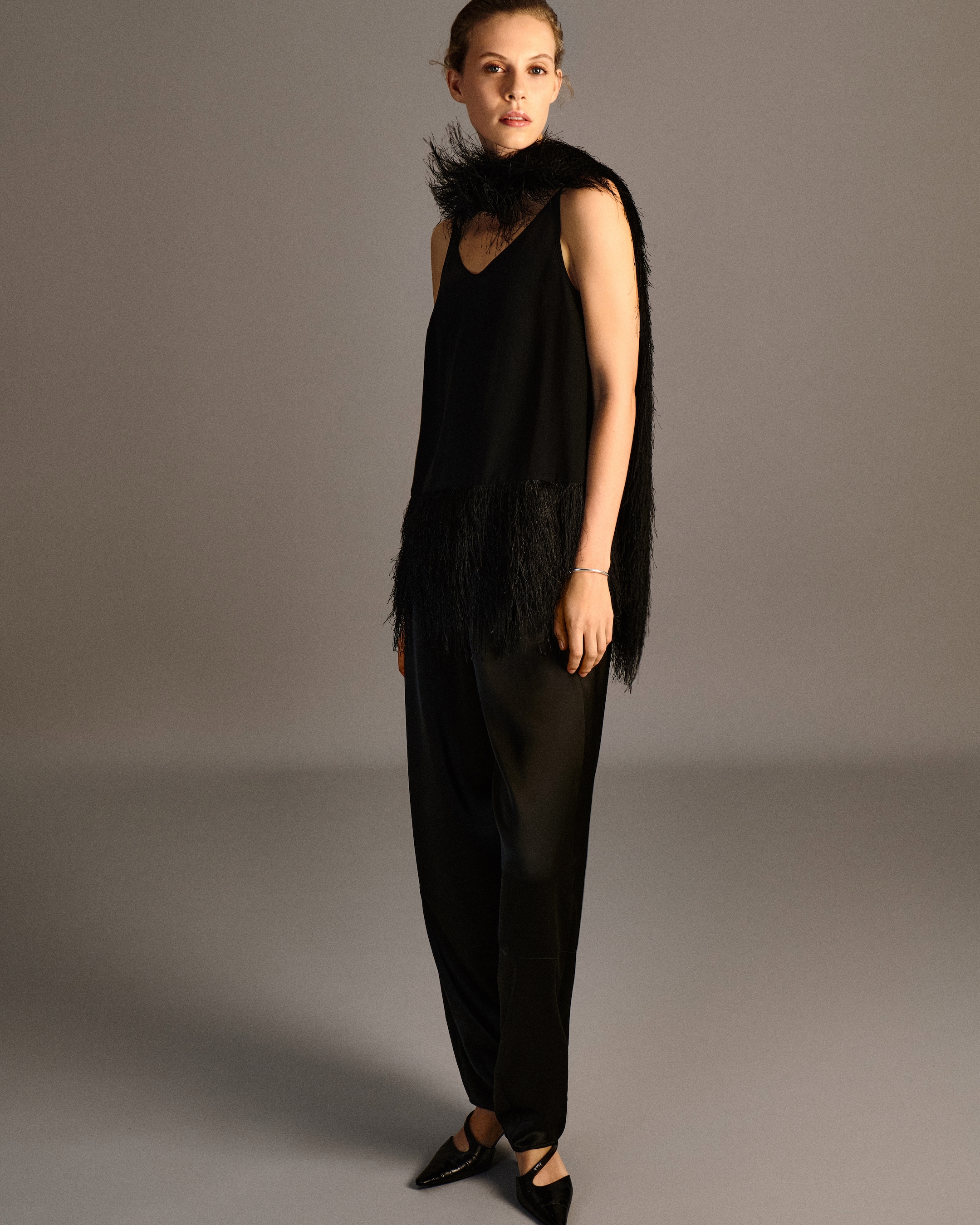 A woman wearing an elegant black outfit with fringe details and a matching scarf, posing against a neutral background.