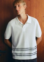Placeholder for: Person with blonde hair, wearing a short-sleeved white shirt with black stripes, stands against a wooden wall, gazing to the side.