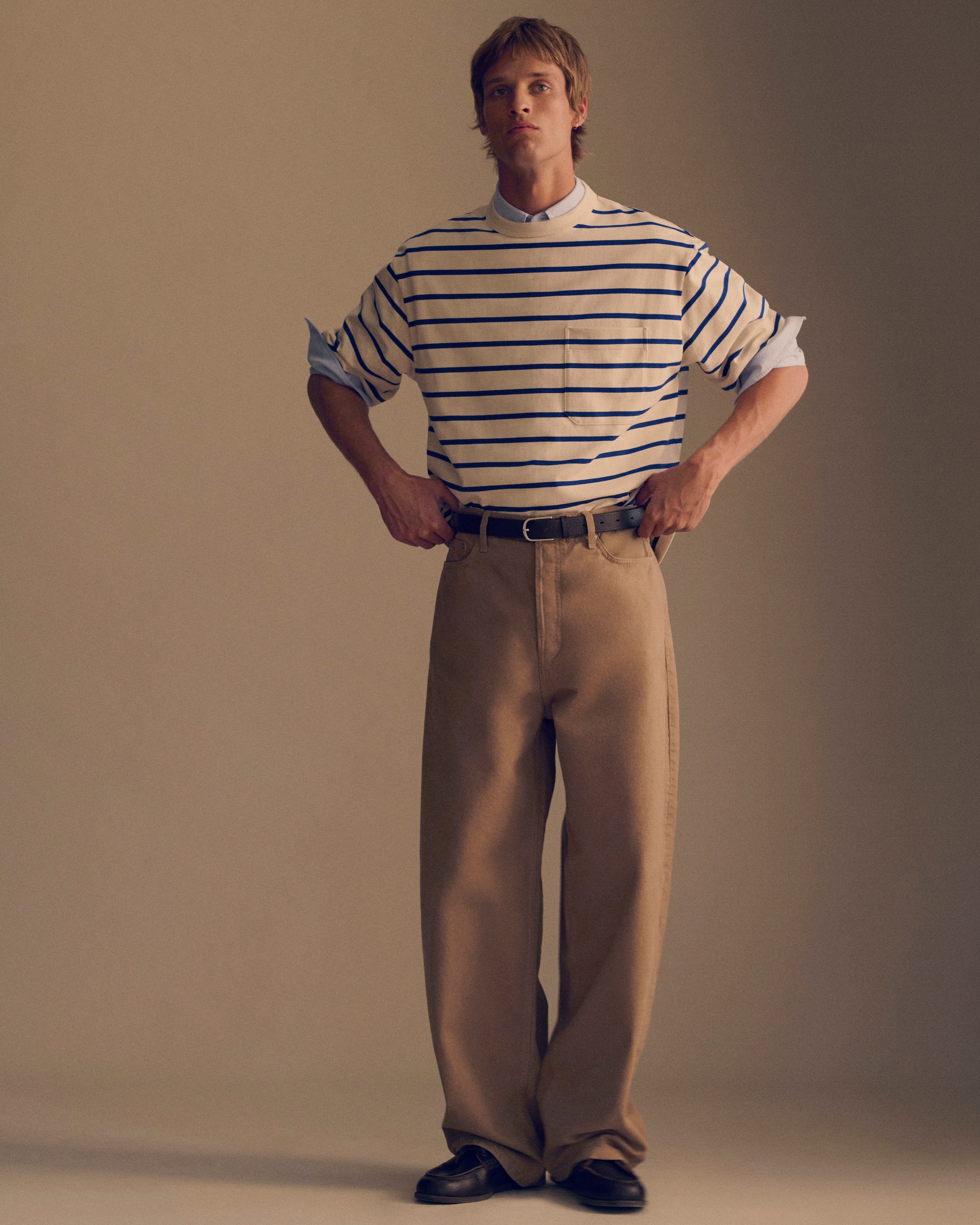 A person stands with hands on hips, wearing a striped shirt, light blue shirt underneath, and wide beige pants, against a plain background.