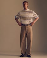 Placeholder for: A person stands with hands on hips, wearing a striped shirt, light blue shirt underneath, and wide beige pants, against a plain background.