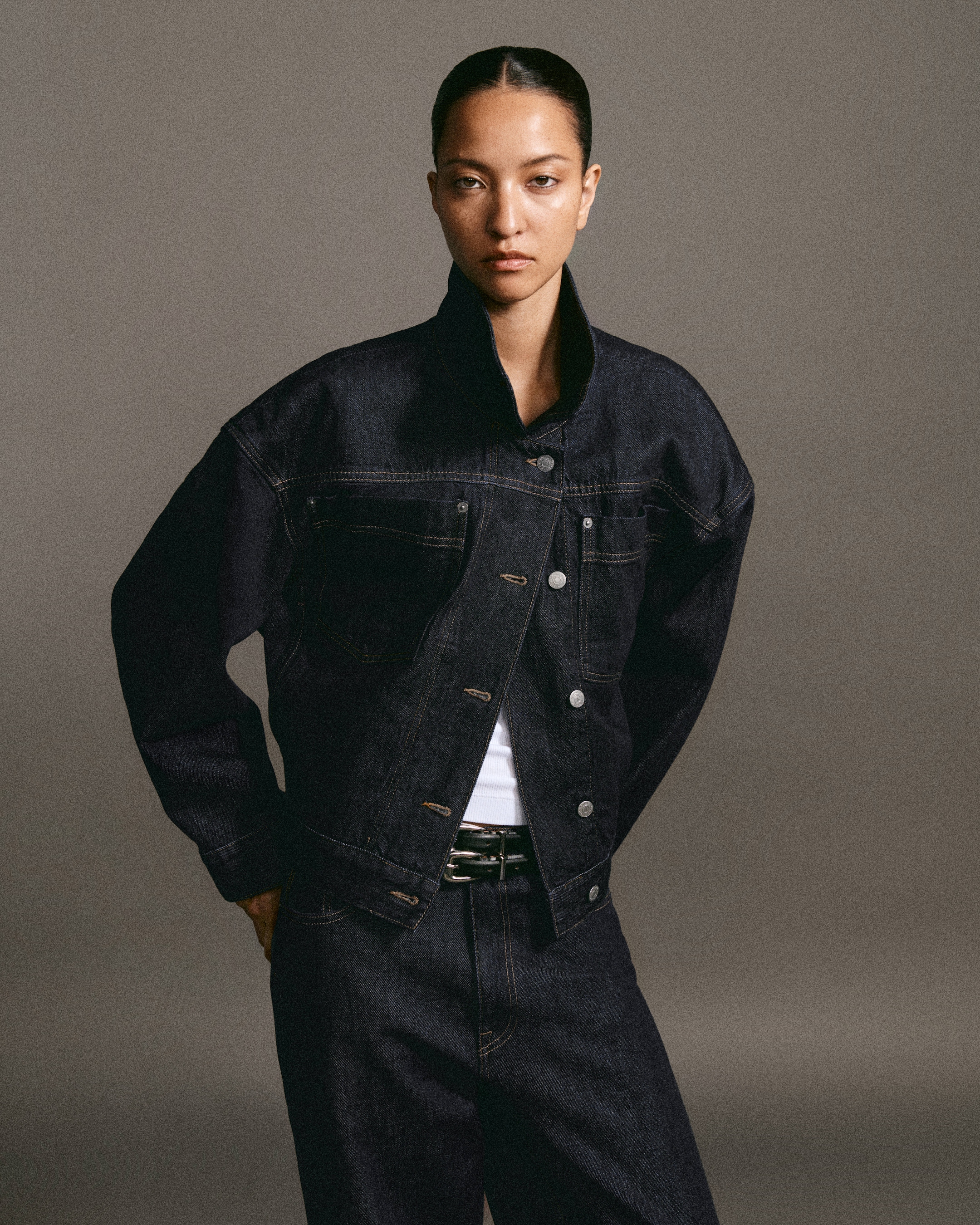 Person wearing a dark denim jacket and pants, posing with a serious expression against a neutral background.