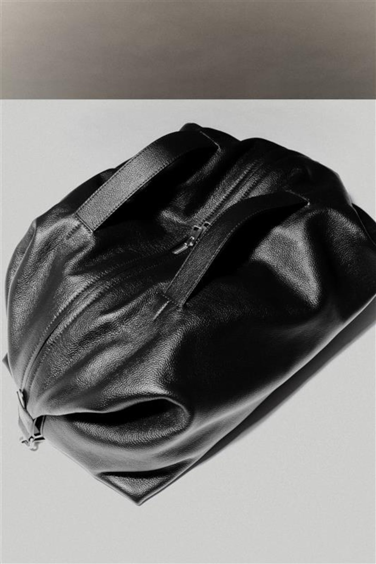 Black leather duffel bag with two handles and a zipper, placed on a light surface, casting a subtle shadow.