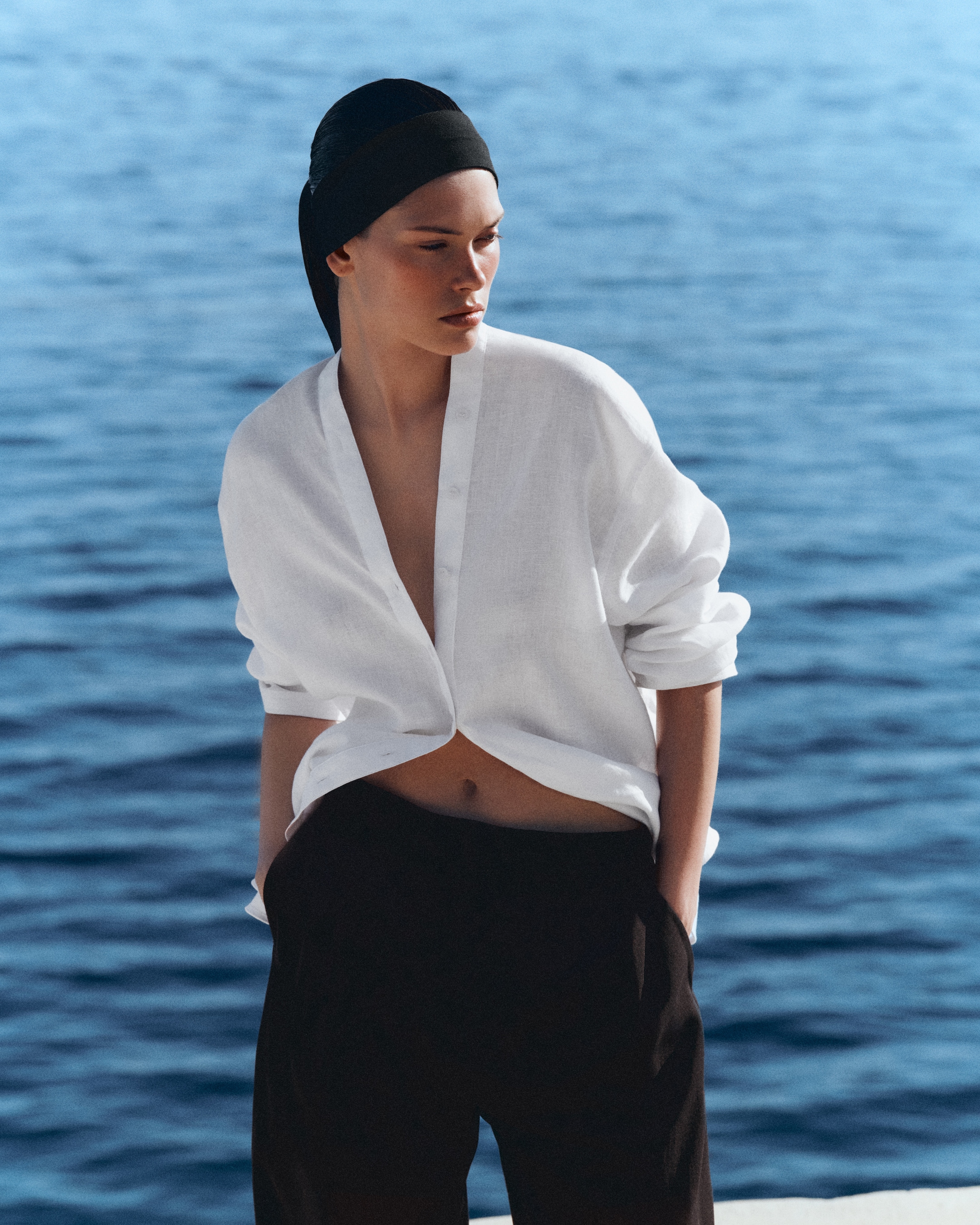 Person in a white shirt and black pants stands by the ocean, gazing thoughtfully. They're wearing a black headscarf.