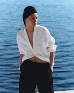 Placeholder for: Person in a white shirt and black pants stands by the ocean, gazing thoughtfully. They're wearing a black headscarf.
