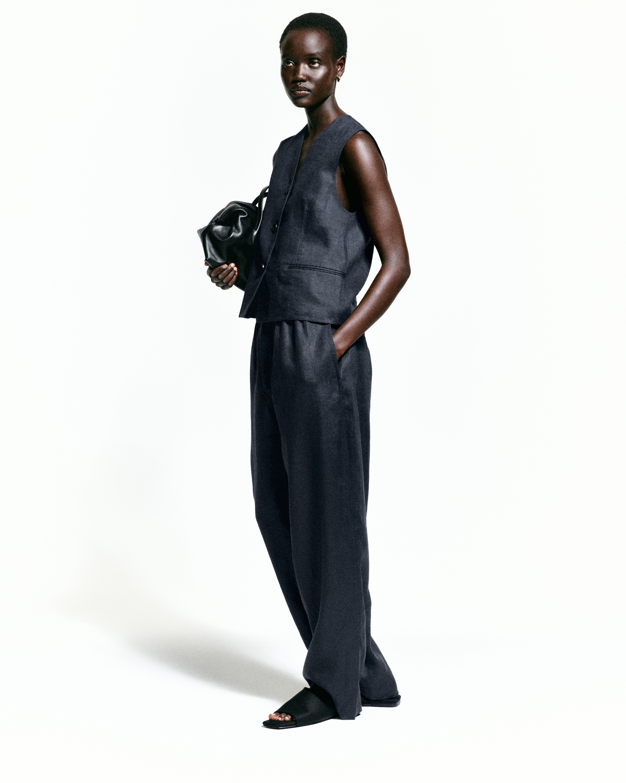 Person in a sleeveless dark outfit, holding a black bag, with one hand in their pocket, standing against a white background.