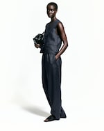 Placeholder for: Person in a sleeveless dark outfit, holding a black bag, with one hand in their pocket, standing against a white background.