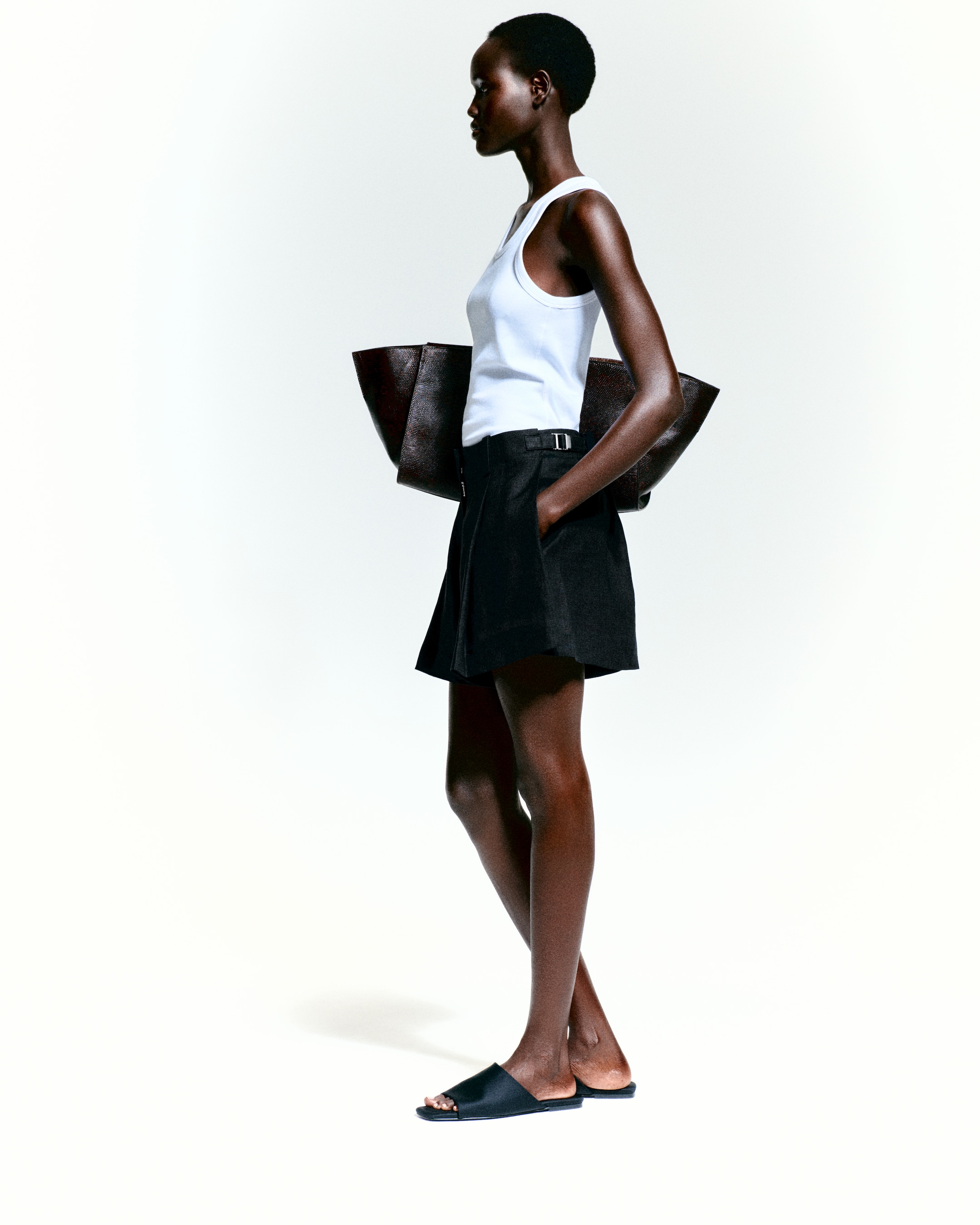 Person in a white tank top, black skirt, and sandals carrying a large dark bag, standing against a light background.
