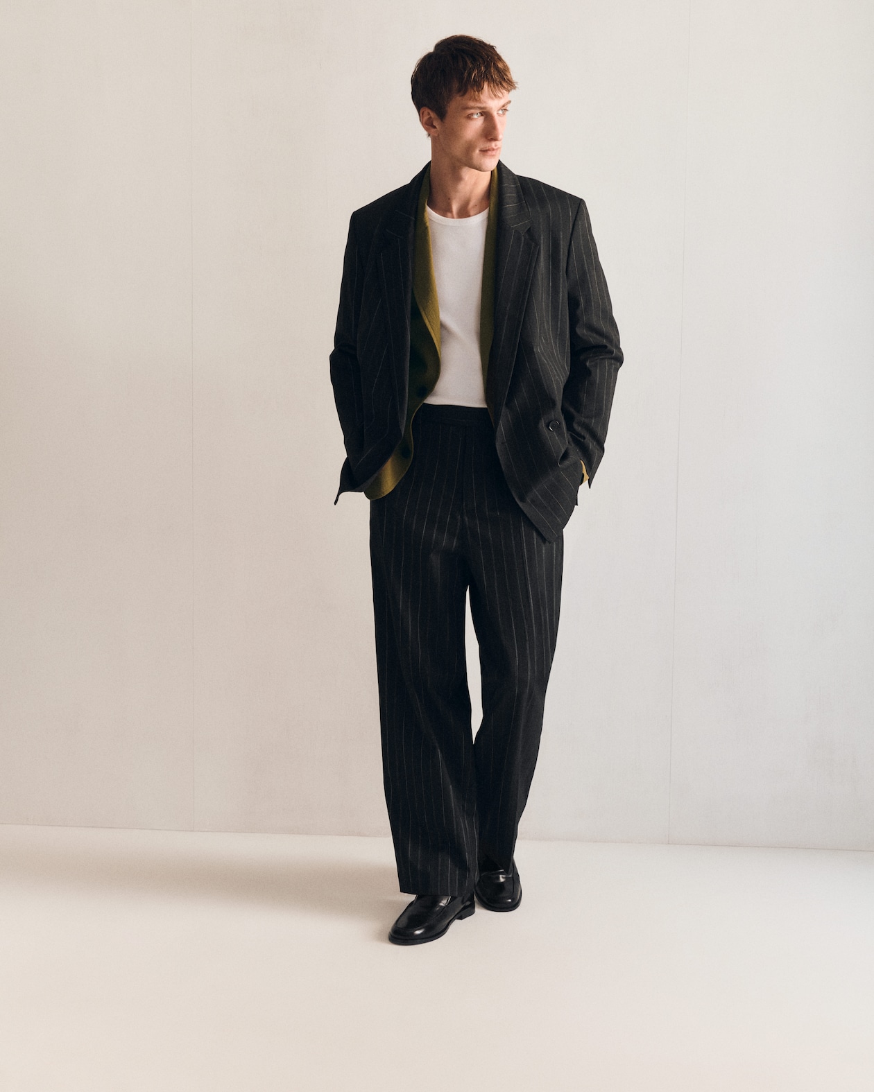 Man in a pinstripe suit over a white shirt stands against a plain background, looking to the side with hands in pockets.