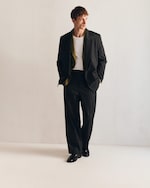 Placeholder for: Man in a pinstripe suit over a white shirt stands against a plain background, looking to the side with hands in pockets.