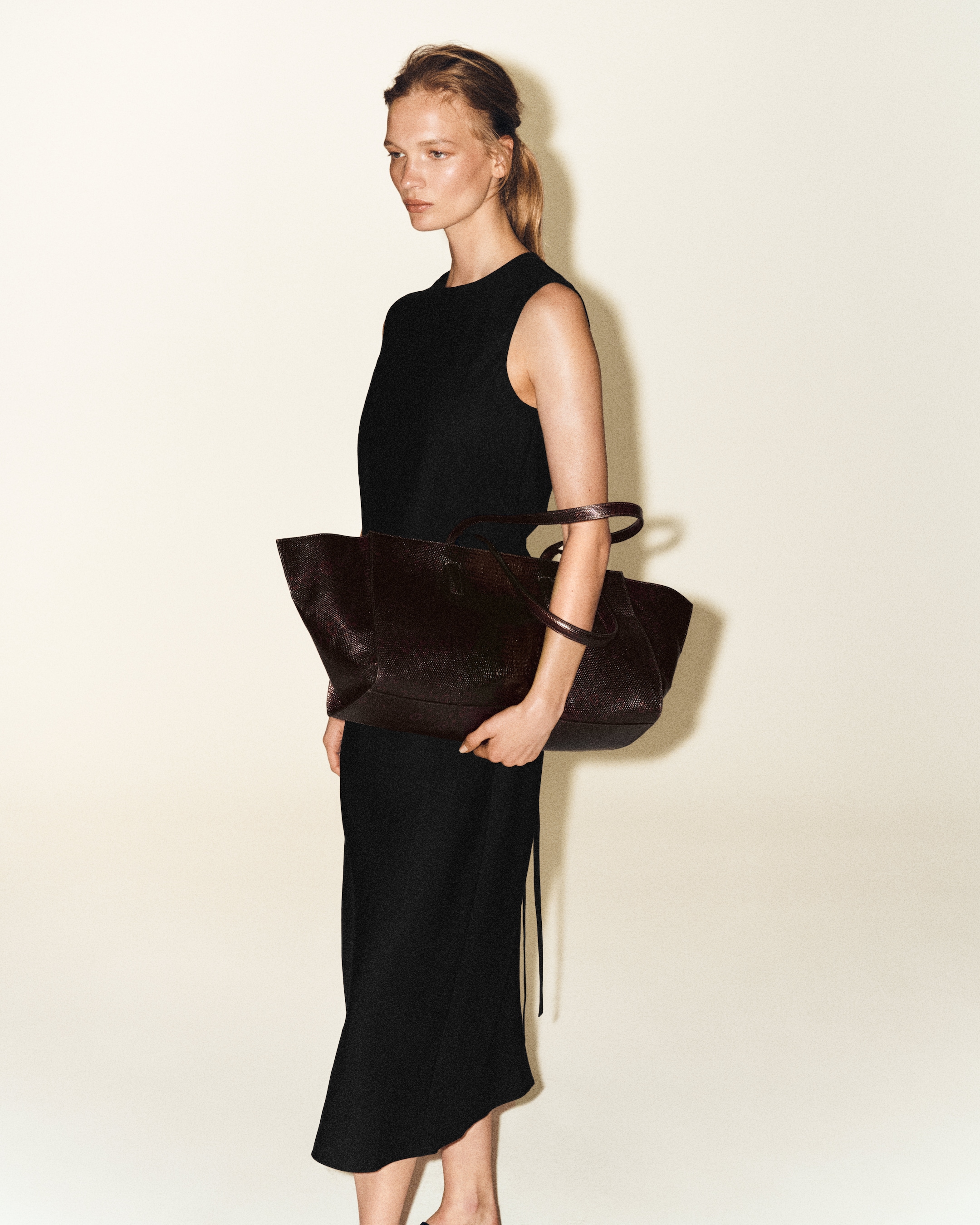 Person in a black sleeveless dress holding a large brown tote bag, standing against a plain light background.