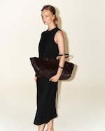 Placeholder for: Person in a black sleeveless dress holding a large brown tote bag, standing against a plain light background.