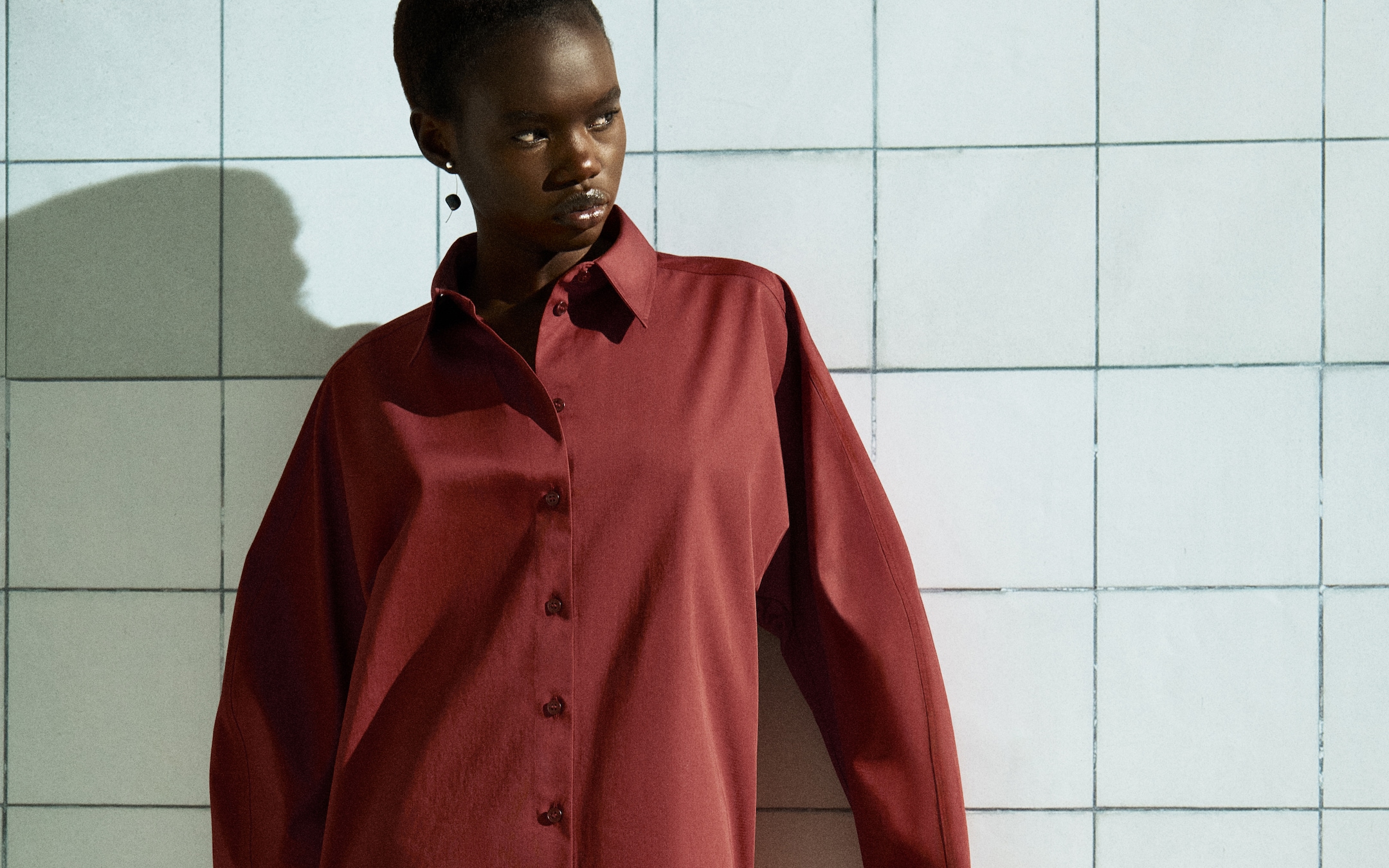 Model wearing a loose-fitting red shirt and dark pants stands against a tiled wall, casting a shadow.