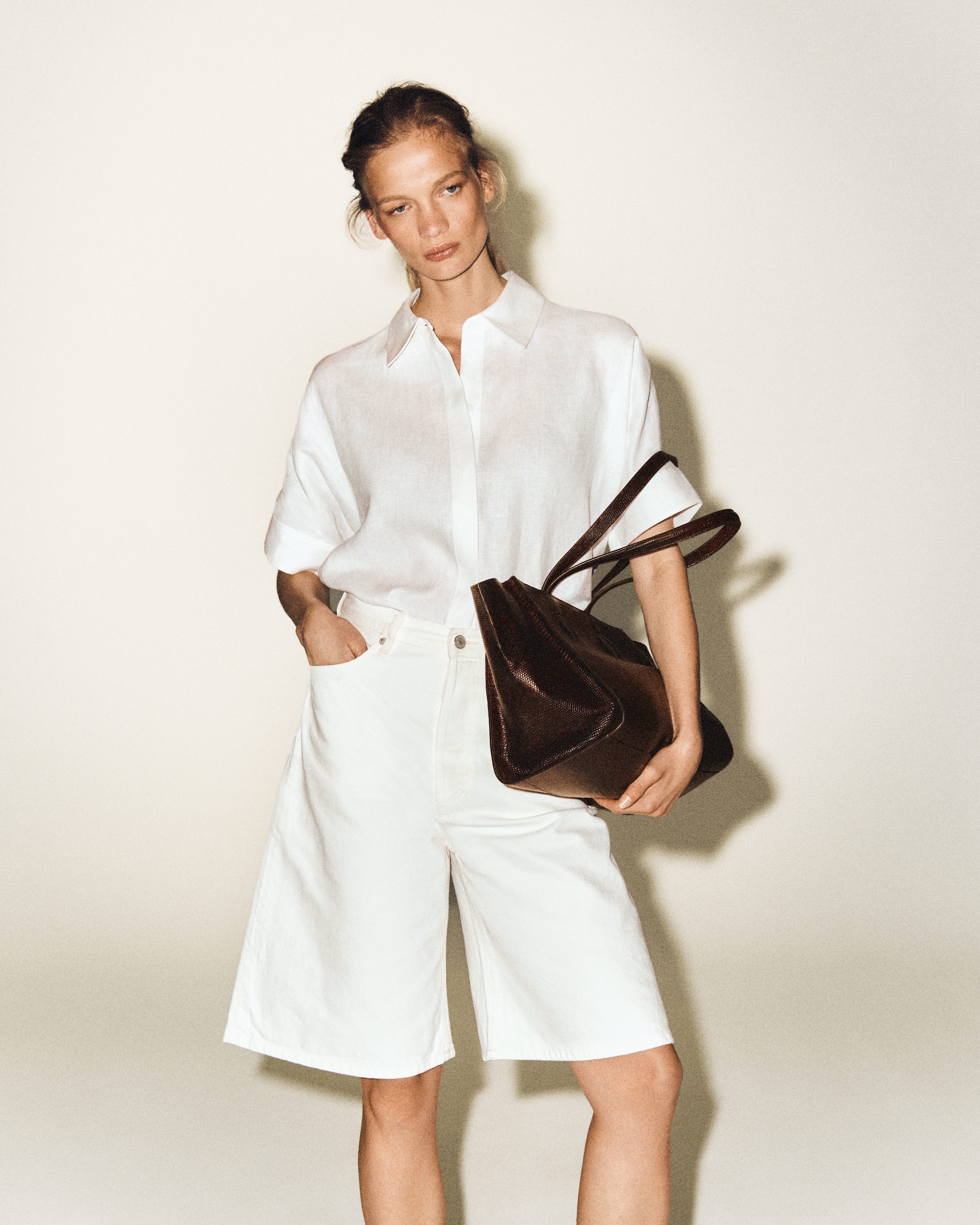 Woman in a white shirt and shorts holding a brown bag, standing against a neutral background.