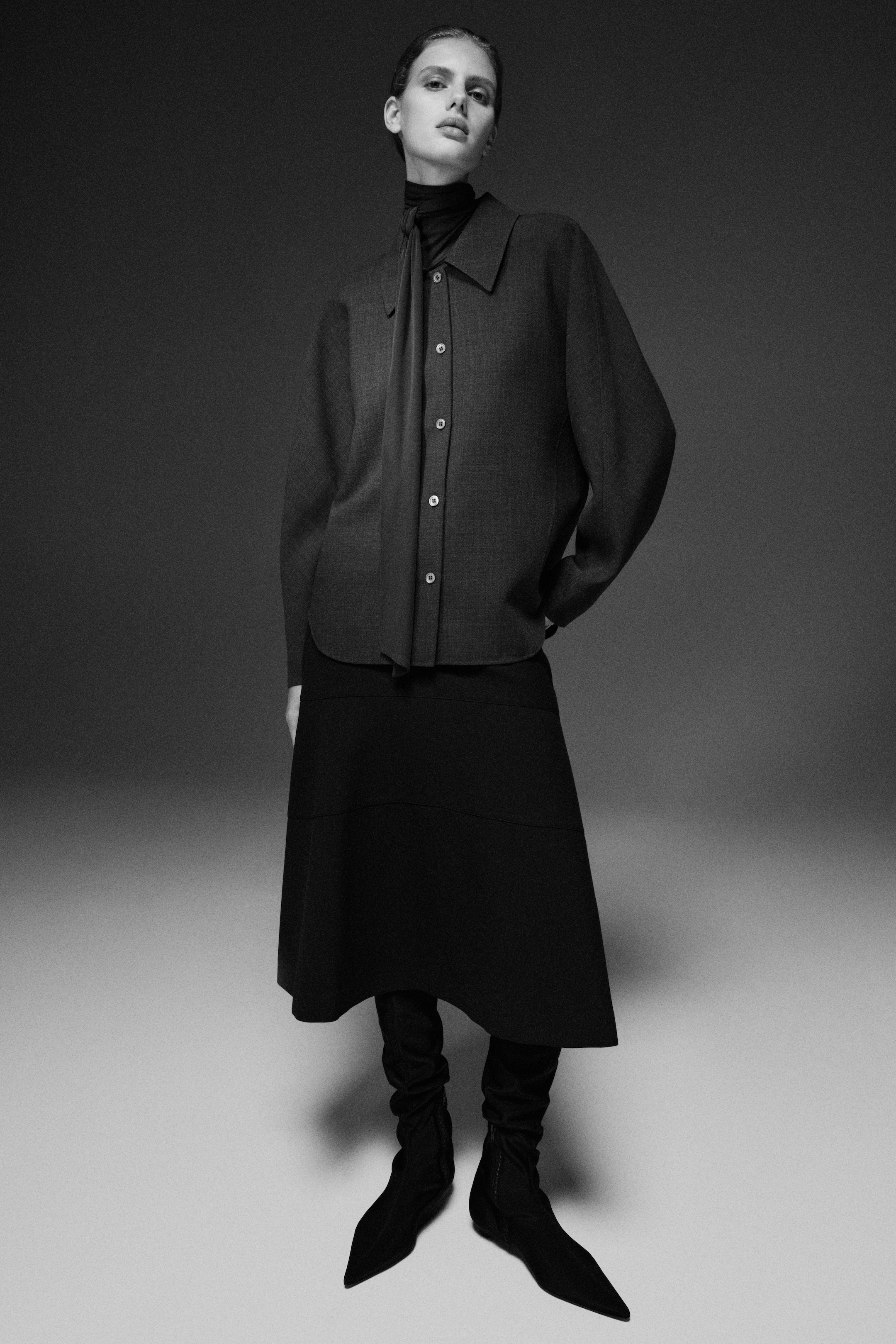 Black-and-white photo of a model in a buttoned blouse with neck scarf, A-line midi skirt and pointed slouchy boots, standing in a studio.