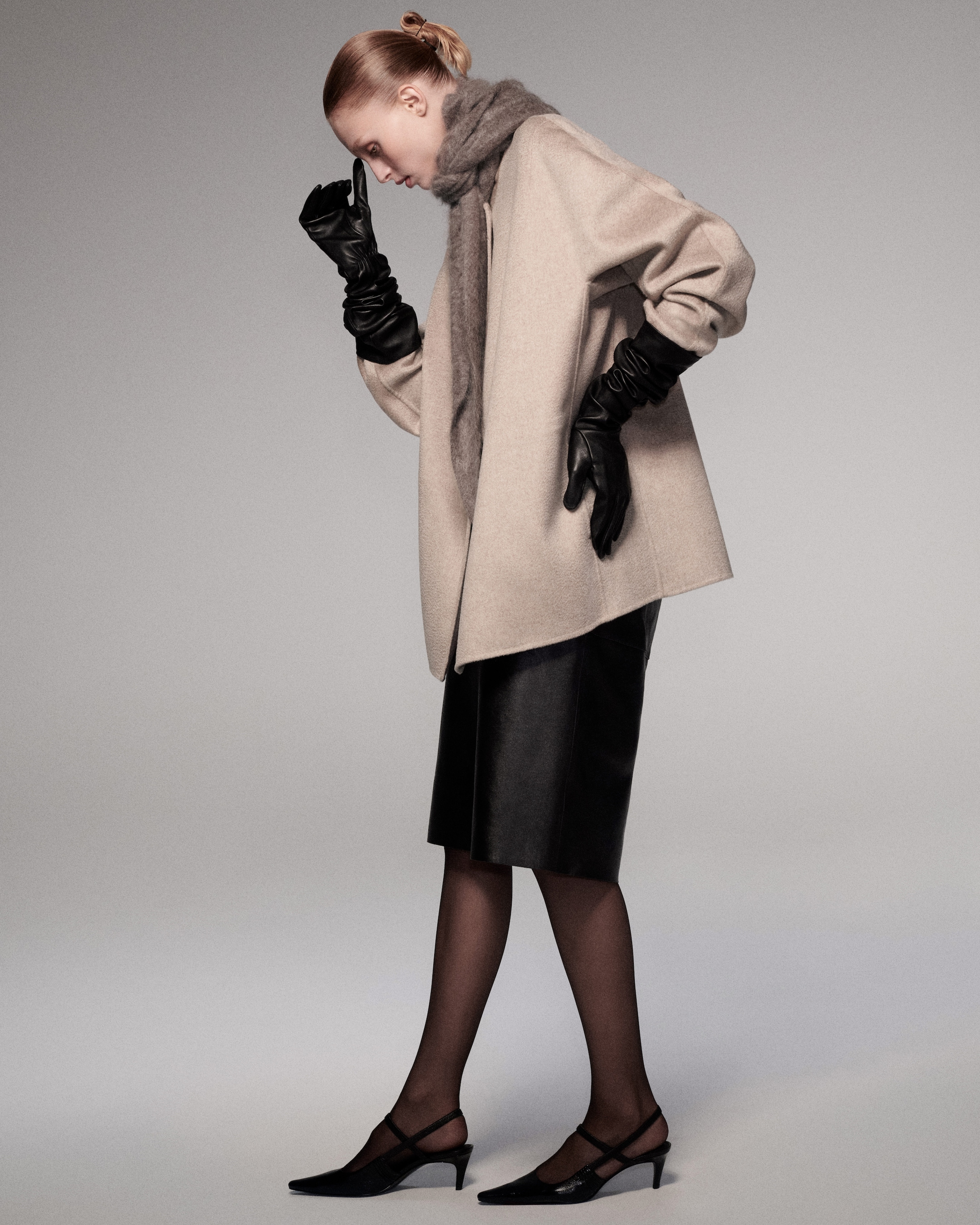 A woman in a beige coat and dark pencil skirt, wearing black gloves and heels, with a grey scarf, standing against a neutral background.