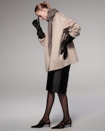 Placeholder for: A woman in a beige coat and dark pencil skirt, wearing black gloves and heels, with a grey scarf, standing against a neutral background.