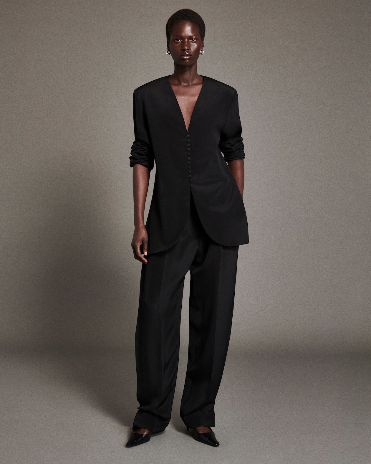 A person wearing an elegant black suit with a deep V-neck, buttoned front, and pants, standing confidently against a neutral background.