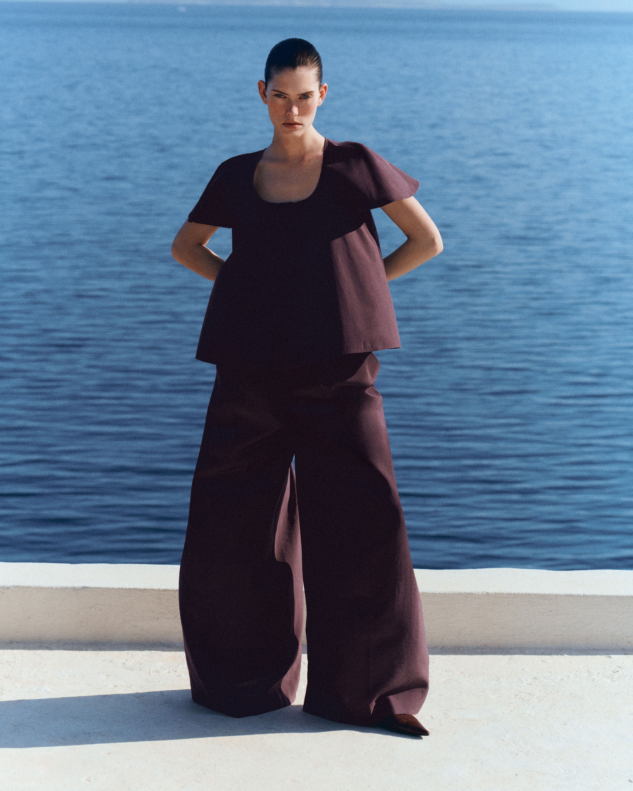 Person in a modern, maroon outfit stands by a waterfront, with hands on hips, against a clear blue sky.