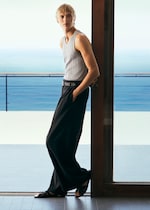 Placeholder for: A person in a gray tank top and black pants leans against a door frame, looking outward with a calm ocean view in the background.