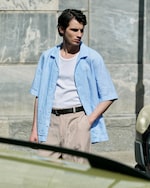 Placeholder for: Man in light blue shirt and beige pants stands near a wall and car, with one hand in pocket, looking to the side.