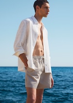 Placeholder for: Man in a white shirt and striped shorts stands by the sea, gazing into the distance on a clear, sunny day.