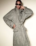 Placeholder for: Person wearing a herringbone-patterned coat and dark sunglasses, posing with one hand on the collar and the other on their hip.