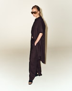 Placeholder for: Person wearing oversized dark outfit, large sunglasses, and open-toed shoes stands against a plain background, hands in pockets.